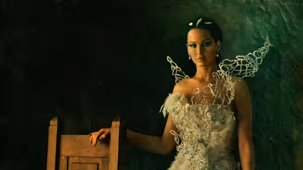 Woman in an elaborate white feathered gown standing beside a wooden chair