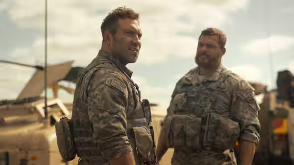 81 (Alan Ritchson) standing beside another soldier near military vehicles outdoors