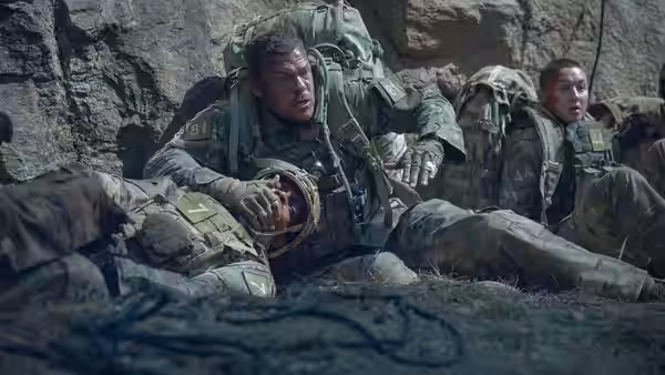 81 (Alan Ritchson) resting against rocks with another soldier in a rocky area