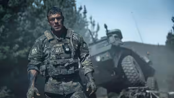 81 (Alan Ritchson) walking toward the camera past a damaged armored vehicle