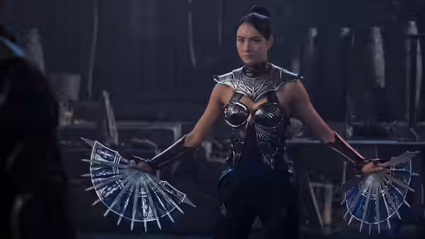 Kitana (Adeline Rudolph) holding two open bladed fans in a dim stone chamber
