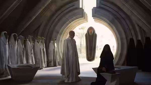 A man in pale robes addresses kneeling figures in a vaulted hall