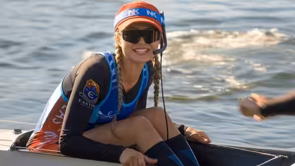 Woman in rowing uniform with headset smiles while seated by water