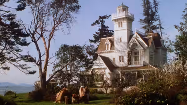 Two figures seated on the lawn before a large white Victorian house with a tower