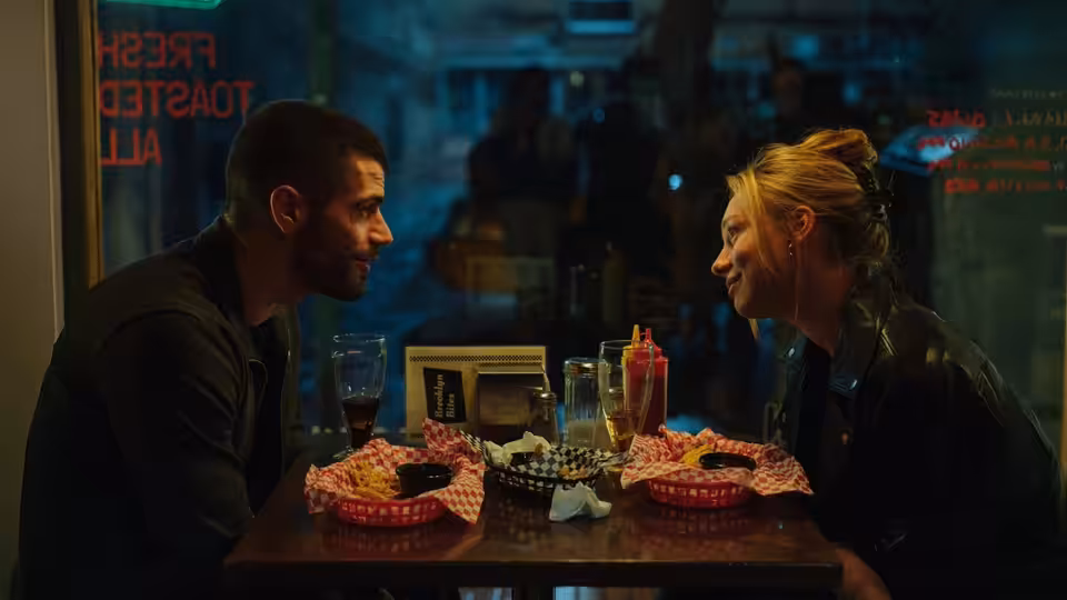 a man and woman face each other across a diner table laden with burgers and drinks