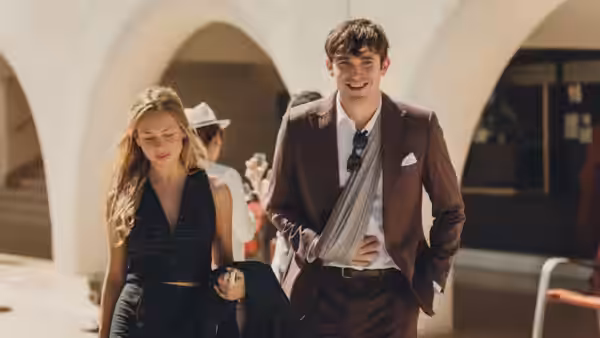 a smiling man in a brown suit walks with a woman through an arched courtyard