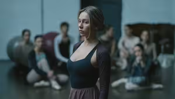 a woman in a leotard stands in the foreground of a dance studio with others seated behind