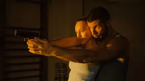 a man and woman together aim a handgun forward in a dimly lit interior