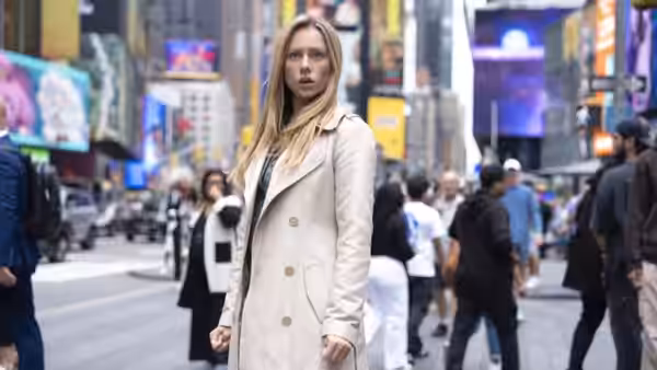 a blonde woman in a beige coat stands amid crowds and billboards in a busy city square