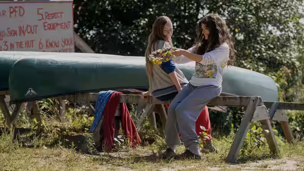 A girl hands flowers to another seated by green canoes outdoors