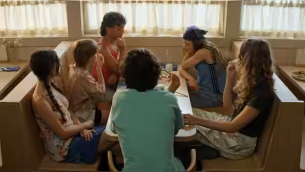Six teenagers sit around a table inside a wooden cabin playing cards