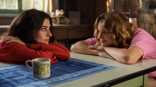 Two young women lean on elbows at a table facing each other