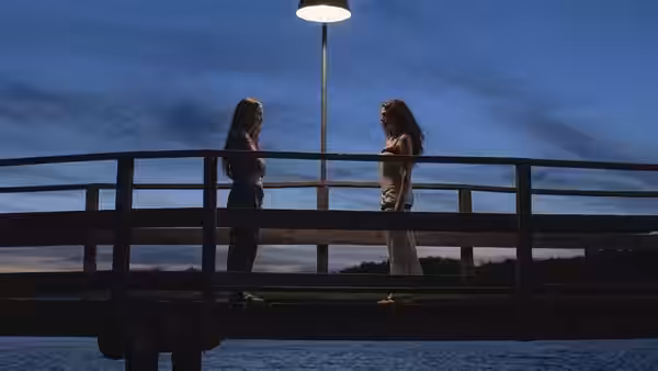 Two women stand facing each other on a wooden bridge at dusk