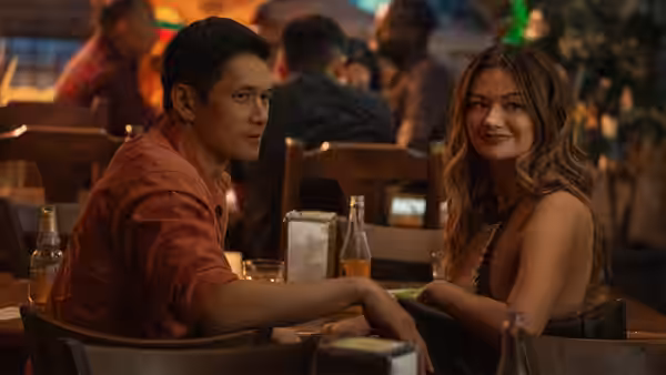 A man in an orange shirt leans on a table while a woman smiles beside him