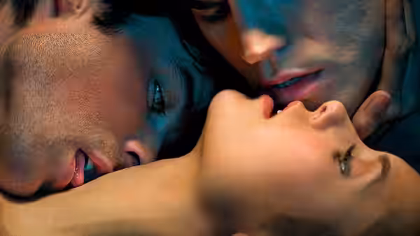 Two men lean close over a woman lying down in dim blue light