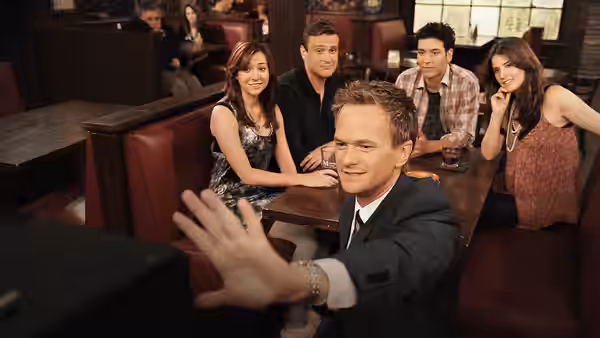 Barney waving at the camera while Lily, Ted, Marshall and Robin sit in a booth
