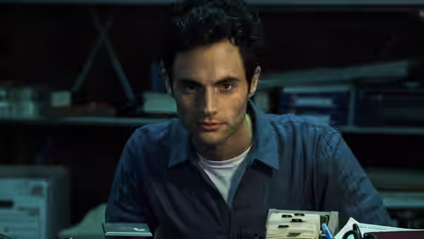 Joe Goldberg (Penn Badgley) seated at a cluttered desk