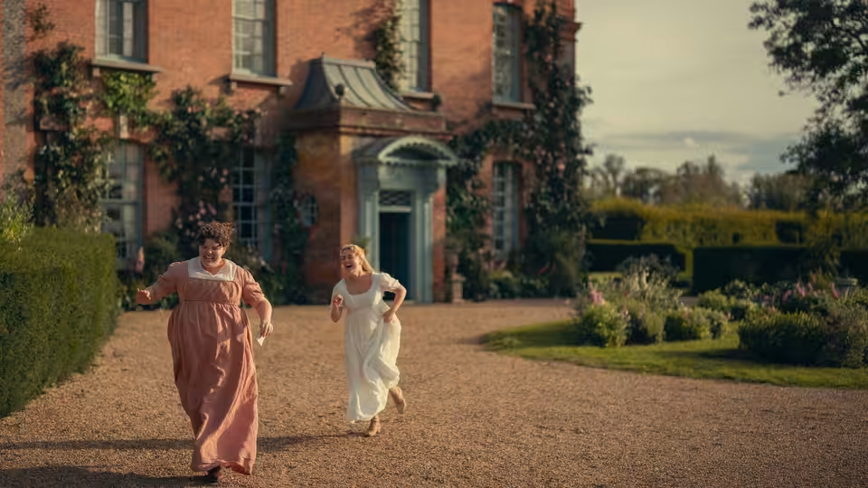Two women in period dresses running on a gravel drive before a brick manor