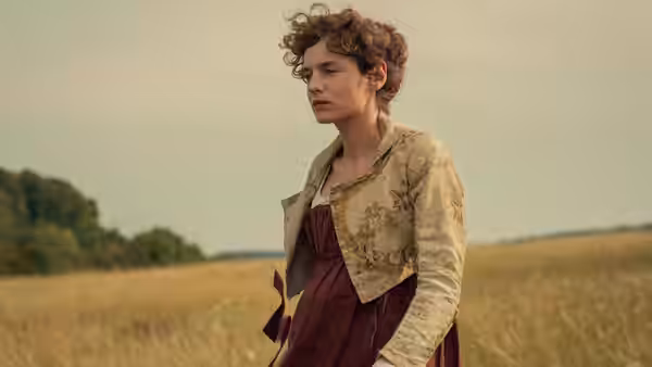 Young woman with curly hair standing in an open grassy field wearing Regency attire