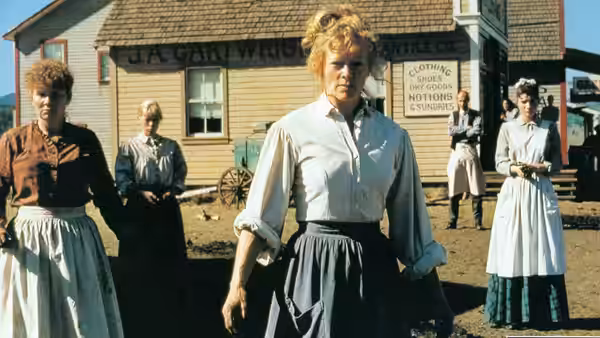 women in long dresses standing outside wooden buildings on a dirt street