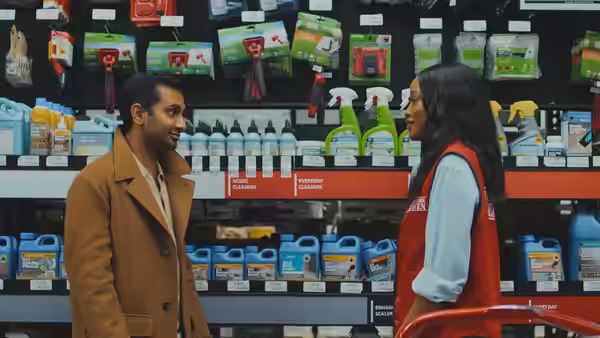 Aziz Ansari speaking with a woman in a red vest inside a hardware store aisle