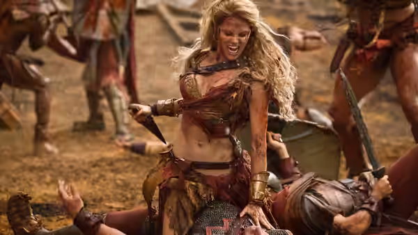 A bloodied female warrior stands victorious over fallen enemies in the arena