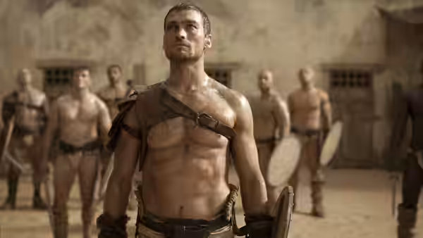 Spartacus (Andy Whitfield) stands shirtless and bloodied in a training yard with gladiators
