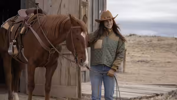 Woman in cowboy hat holding reins of brown horse outside wooden barn