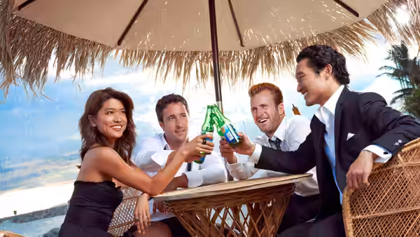 Four friends toast with green bottles at a beachside table under a thatched umbrella