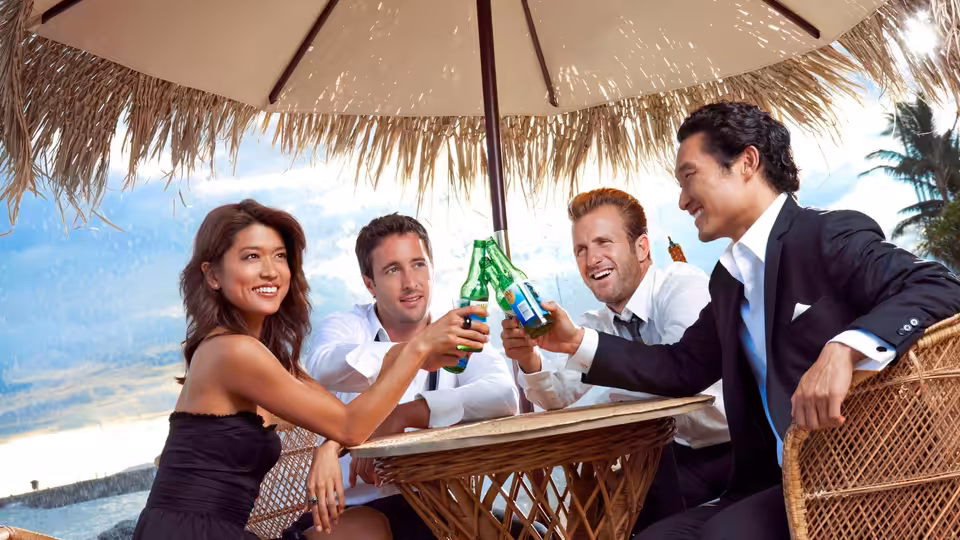 Four friends toast with green bottles at a beachside table under a thatched umbrella