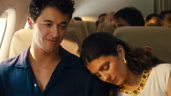 Alex (Tom Blyth) sitting beside Poppy (Emily Bader) resting on his shoulder on a plane