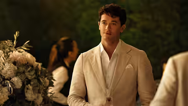 Alex (Tom Blyth) in a white suit standing at an outdoor formal event