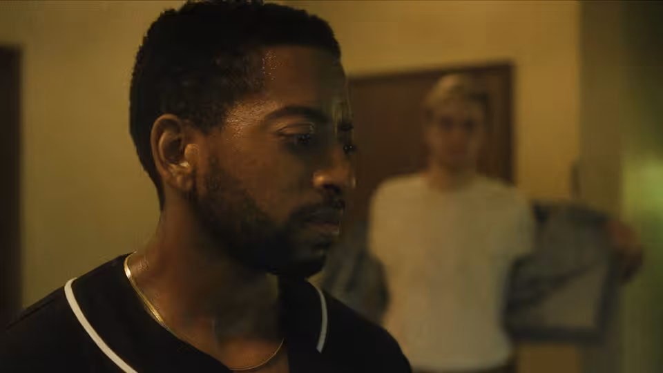 A bearded man in a black shirt looks sideways with a person in the background