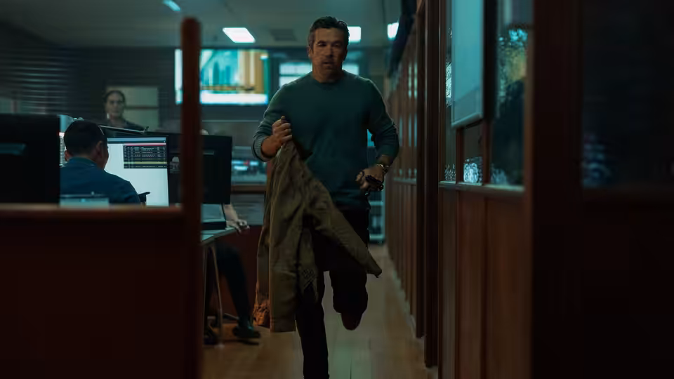 A man runs down an office hallway holding a jacket and pistol