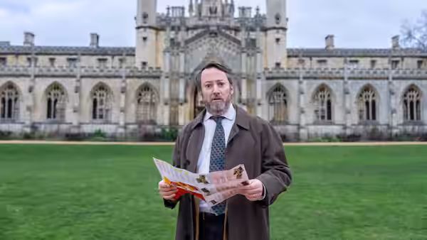 John Taylor (David Mitchell) holding papers on a lawn before a building