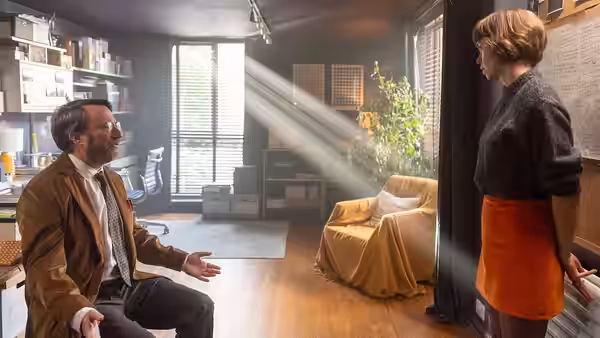John Taylor (David Mitchell) seated and gesturing to a woman indoors