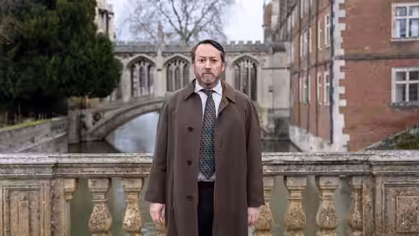 John Taylor (David Mitchell) standing on a stone bridge by water