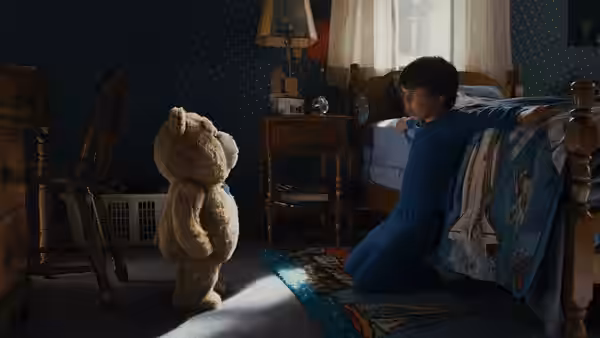 John sits beside his bed facing Ted in a dim bedroom