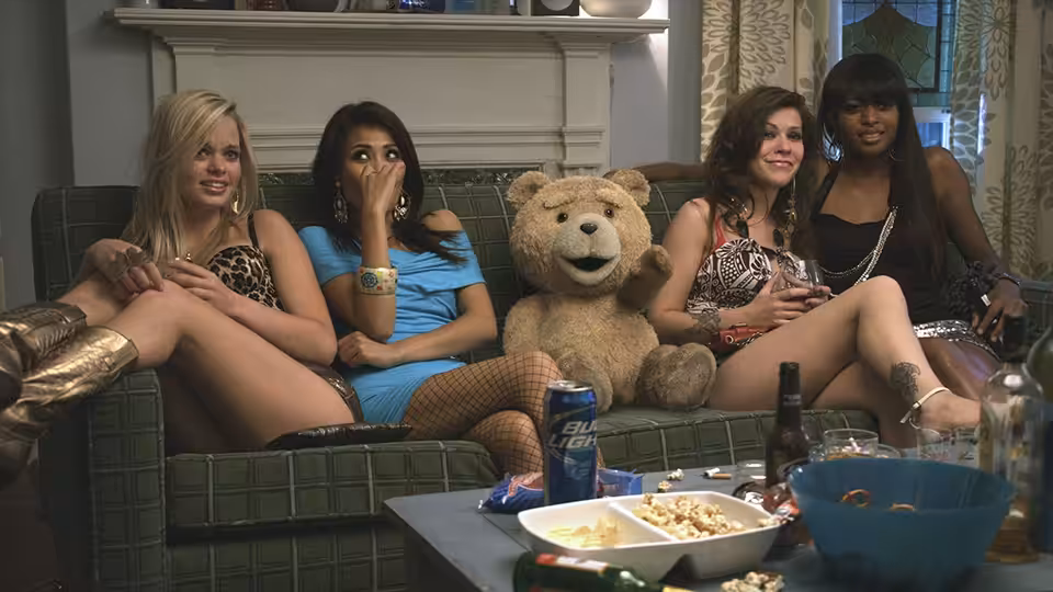 Women lounge with Ted on a living-room couch amid drinks and party snacks