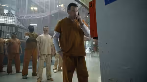 Man in orange jumpsuit speaking on phone in prison with other inmates nearby