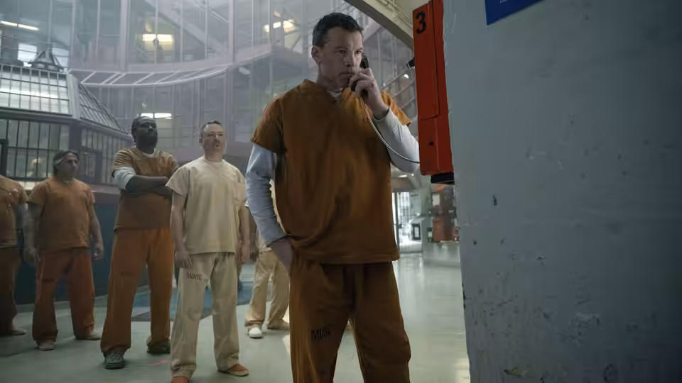 Man in orange jumpsuit speaking on phone in prison with other inmates nearby
