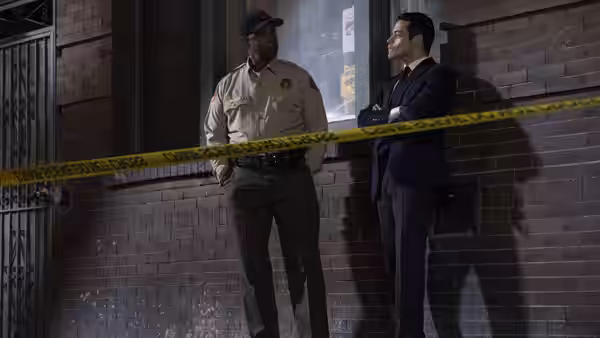 A police officer and Rami Malek stand behind yellow crime scene tape