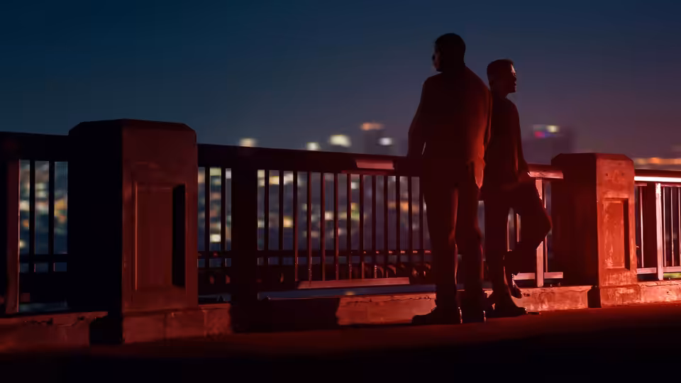 Two men stand on a bridge overlooking a city glowing at night