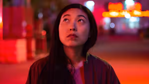 Billi (Awkwafina) gazes upward on a neon-lit street at night