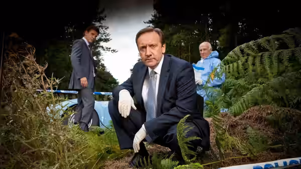 Detective crouching at a wooded crime scene with officers nearby