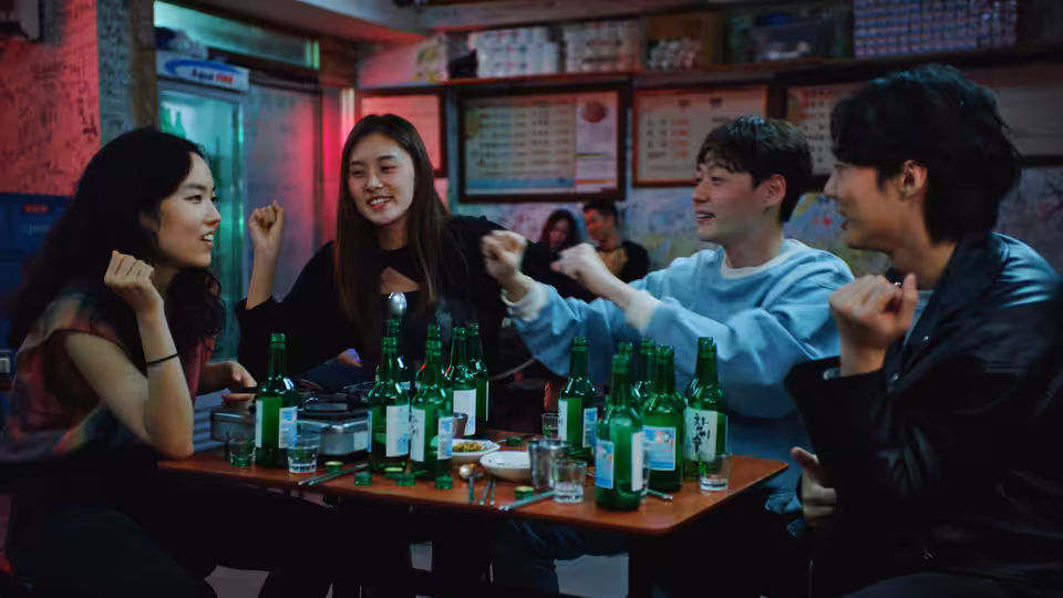 Freddie (Ji-min Park) and three friends laugh and gesture around a table of green bottles