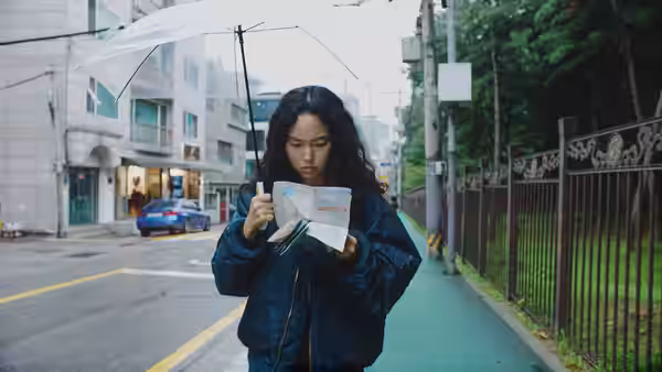 Freddie (Ji-min Park) studies a map under a clear umbrella on a city sidewalk