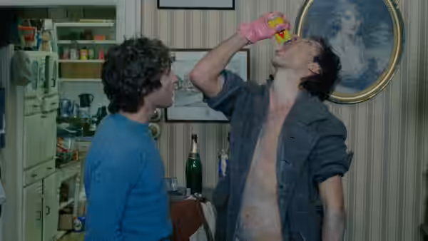 Withnail (Richard E. Grant) drinking from a bottle in a kitchen as Marwood (Paul McGann) watches