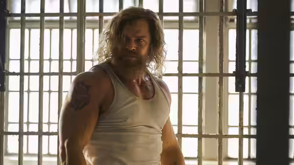 Long-haired bearded man in tank top standing before prison cell bars