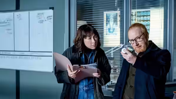 DI Max Arnold (Adrian Scarborough) pointing a remote as Astrid Fischer reviews papers